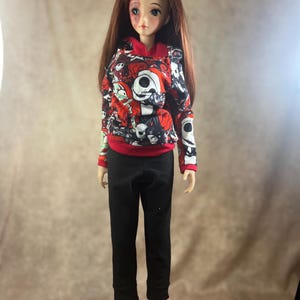 Puede incluir: Una mu&ntilde;eca con pelo largo casta&ntilde;o que lleva una sudadera con capucha roja y negra con un dise&ntilde;o de Jack Skellington, pantalones negros y zapatos negros. La mu&ntilde;eca tiene un hematoma en la frente. La sudadera tiene pu&ntilde;os rojos y una capucha roja.