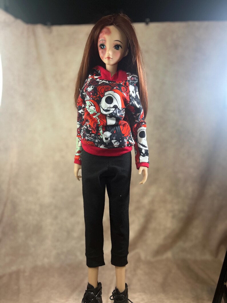 Puede incluir: Una mu&ntilde;eca con pelo largo casta&ntilde;o que lleva una sudadera con capucha roja y negra con un dise&ntilde;o de Jack Skellington, pantalones negros y zapatos negros. La mu&ntilde;eca tiene un hematoma en la frente. La sudadera tiene pu&ntilde;os rojos y una capucha roja.