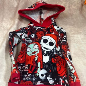 Puede incluir: Una sudadera con capucha con un estampado colorido de Jack Skellington y Sally de Pesadilla antes de Navidad. El dise&ntilde;o incluye rosas rojas, calaveras y otros elementos tem&aacute;ticos. La capucha y los pu&ntilde;os est&aacute;n ribeteados en rojo.