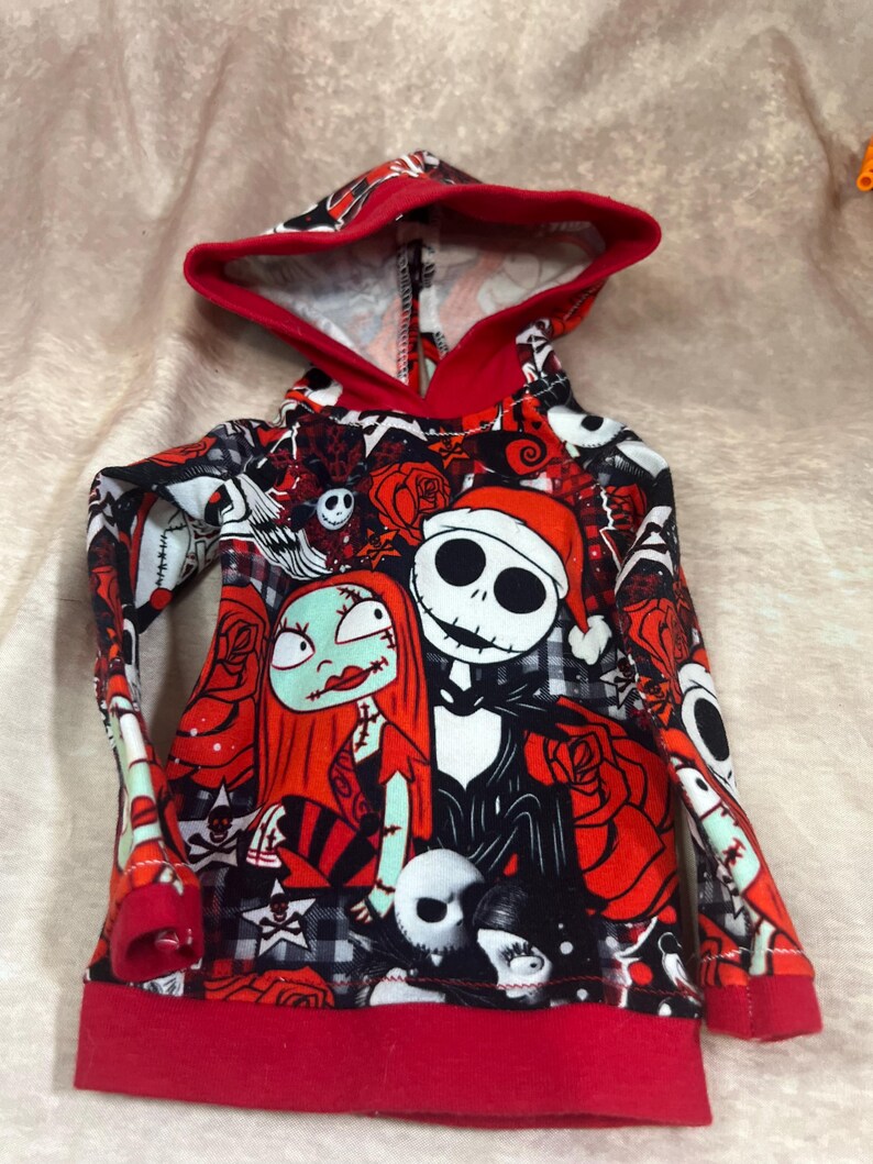 Puede incluir: Una sudadera con capucha con un estampado colorido de Jack Skellington y Sally de Pesadilla antes de Navidad. El dise&ntilde;o incluye rosas rojas, calaveras y otros elementos tem&aacute;ticos. La capucha y los pu&ntilde;os est&aacute;n ribeteados en rojo.