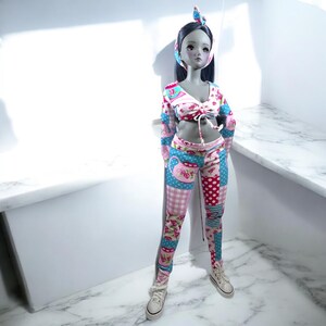 Könnte beinhalten: Eine Puppe, die ein rosafarbenes und blaues, gemustertes Outfit mit einem passenden Stirnband trägt. Das Outfit besteht aus einem bauchfreien Oberteil mit einem Bindedetail und Leggings mit Patchwork-Muster.