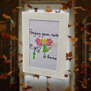 Könnte beinhalten: Eine weiße Karte mit helllila Hintergrund und einer handgezeichneten Illustration einer Fee mit rosa Flügeln und einem grünen Kleid. Der Text "Enjoy your new home" ist in schwarzer Kursivschrift geschrieben.