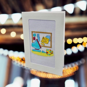 May include: A white card with a light blue, yellow, red, and green cross-stitch design of a mug, a bottle, and a sign that says "Not a Bar".