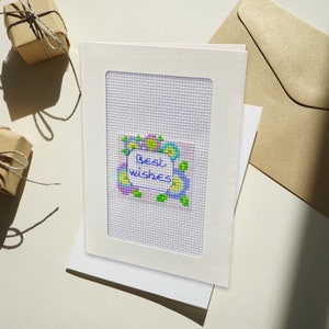 Può includere: Un biglietto bianco con una cornice bianca attorno a un disegno a punto croce di fiori colorati con il testo "Auguri" in blu.