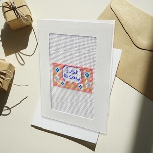 Può includere: Un biglietto bianco con un disegno a punto croce rosa e bianco. Il disegno presenta un rettangolo rosa con fiori bianchi e il testo "Just to say" in blu.