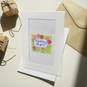 Può includere: Un biglietto bianco con un bordo bianco e uno sfondo giallo con fiori rosa e blu. Il testo "Thinking of you" è scritto in inchiostro blu.