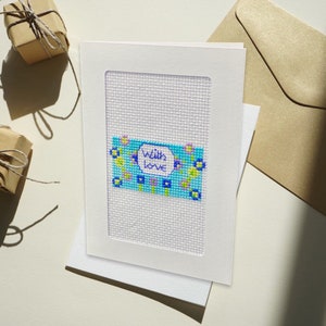 Può includere: Un biglietto bianco con un ricamo a punto croce blu e verde con fiori e il testo "With love".