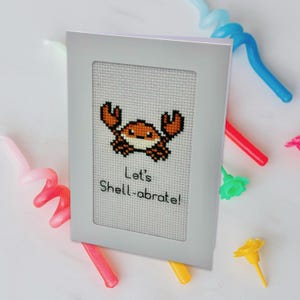 May include: A white card with a white frame around a cross-stitch design of a brown crab with the text "Let's Shell-abrate!"