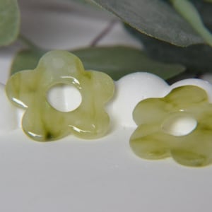 May include: Two flower-shaped beads with a central hole. The beads are a translucent, pale green color with darker green veining. They are set against a white, textured surface with green foliage in the background. These beads are suitable for jewelry making.