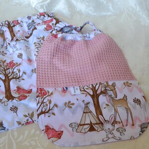 May include: Two drawstring bags with a woodland theme. One bag is white with a repeating pattern of deer, trees, and flowers. The other bag has a pink waffle-textured top and a similar woodland pattern on the bottom.