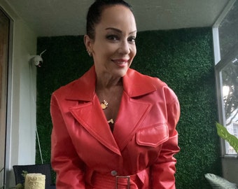May include: A woman wearing a red leather jacket and matching shorts. The jacket has a belt and a pocket on the left side. The woman is smiling and looking at the camera.