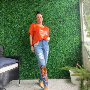 Pode incluir: Uma mulher vestindo uma blusa laranja brilhante com lantejoulas e jeans azul com um design de tigre. Ela está em pé em frente a uma parede verde.