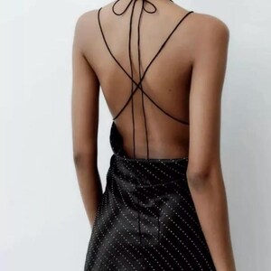 Puede incluir: Un vestido negro de sat&eacute;n corto con estampado de lunares blancos. El vestido tiene la espalda baja con tirantes finos que se atan en el cuello.