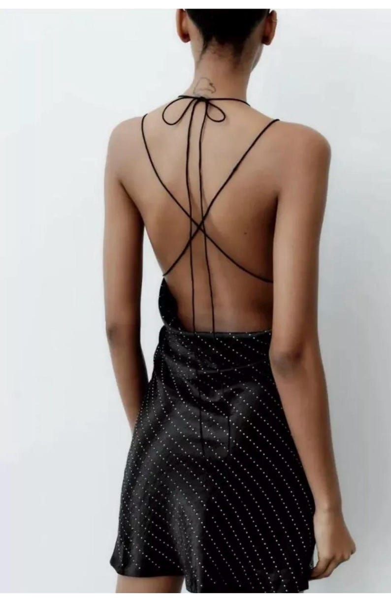 Puede incluir: Un vestido negro de sat&eacute;n corto con estampado de lunares blancos. El vestido tiene la espalda baja con tirantes finos que se atan en el cuello.
