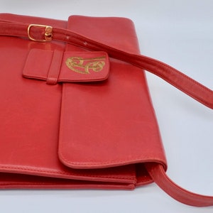 May include: A red leather shoulder bag with a gold-colored buckle and a flap with a gold-colored design.