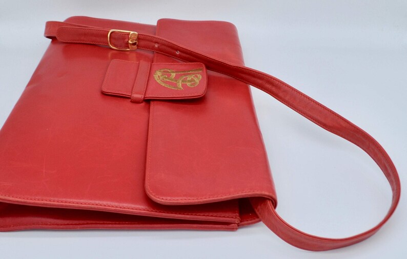 May include: A red leather shoulder bag with a gold-colored buckle and a flap with a gold-colored design.
