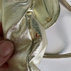 May include: A gold-colored leather handbag with a small, round, gold-colored button on the flap. The bag has a strap and is slightly wrinkled.