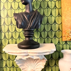 May include: A black bust of a woman with a bun hairstyle, atop a decorative shelf. The shelf is white and shell-shaped, mounted against a green and white patterned wall. A white vase with floral details is visible.