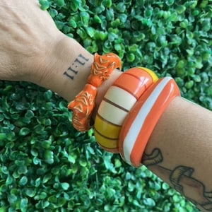 Puede incluir: Un conjunto de tres pulseras de plástico de colores en naranja, amarillo y blanco. Las pulseras están apiladas en la muñeca de una persona.