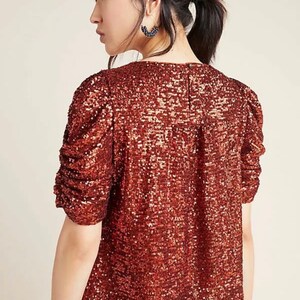 Pode incluir: Uma blusa de lantejoulas cor ferrugem com decote redondo e mangas curtas bufantes.
