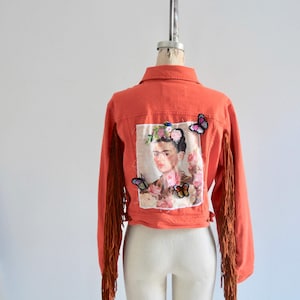 May include: A coral colored denim jacket with brown fringe on the sleeves. The back of the jacket features a printed image of Frida Kahlo with butterflies.