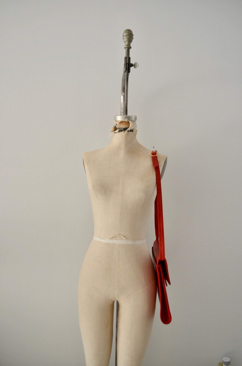 May include: A red leather shoulder bag with a long strap hanging on a white dress form.