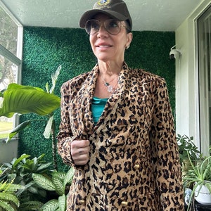 May include: A woman wearing a brown leopard print blazer with gold buttons, a teal tank top, and a brown baseball cap with a gold emblem. She is standing in front of a green wall.