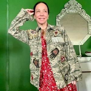 Può includere: Una giacca mimetica con un vestito floreale rosso sotto. La giacca è decorata con cuori ricamati, mani e altri abbellimenti.