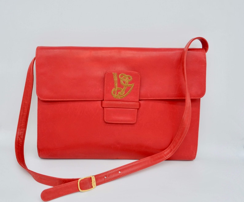 May include: A red leather shoulder bag with a gold embroidered logo. The bag has a flap closure and a long adjustable strap.