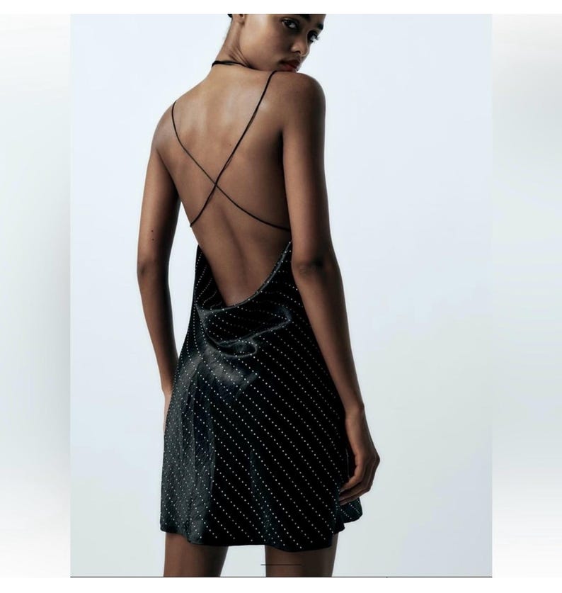 Puede incluir: Un vestido corto de sat&eacute;n negro con estampado de lunares blancos y tirantes cruzados en la espalda.