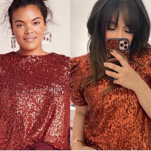 Pode incluir: Duas mulheres vestindo uma blusa de lantejoulas cor ferrugem com mangas curtas e um ombro levemente bufante. A blusa tem um decote redondo.