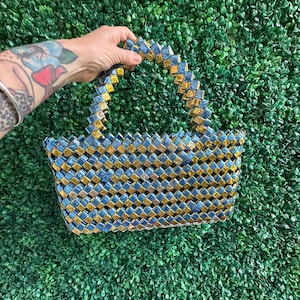 Puede incluir: Un bolso tejido azul y dorado con un patrón cuadrado. El bolso tiene un asa superior y está hecho de un material brillante.