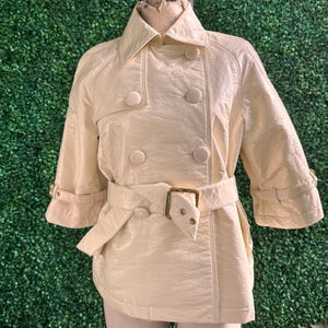 May include: Cream-colored trench coat with a double-breasted design, featuring large round buttons and a matching belt with a gold-colored buckle. The coat has three-quarter sleeves and is displayed on a mannequin against a green backdrop.