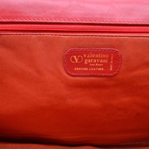 May include: Close-up of a red leather purse interior with a gold label that reads "Valentino Garavani, Made in Italy, Genuine Leather".