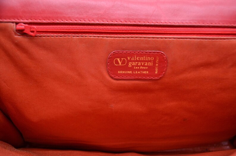 May include: Close-up of a red leather purse interior with a gold label that reads "Valentino Garavani, Made in Italy, Genuine Leather".