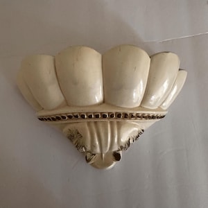 May include: Off-white, shell-shaped wall sconce. The decorative piece features a scalloped top, a detailed lower section, and a small, rounded base. The sconce is made of a solid material and has a glossy finish.