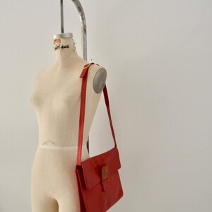 May include: A red leather shoulder bag with a flap closure and a gold buckle. The bag is hanging on a dress form.