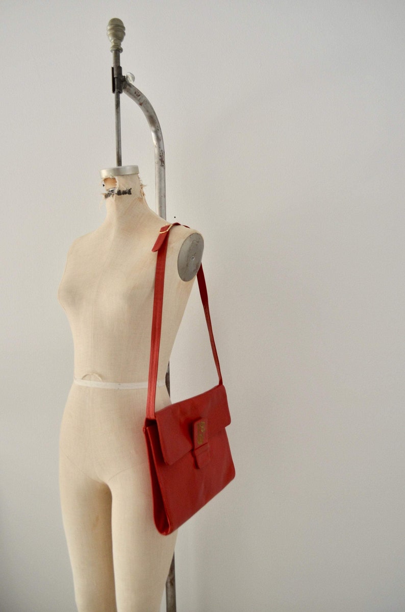 May include: A red leather shoulder bag with a flap closure and a gold buckle. The bag is hanging on a dress form.