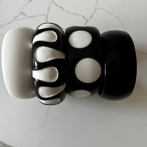 May include: Four stacked, chunky bangles in a row. The first is solid white, the second is black with white teardrop shapes, the third is black with white polka dots, and the last is solid black. The bangles are made of a glossy material.