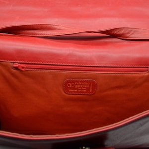 May include: Red leather handbag with a flap closure and a shoulder strap. The inside of the bag is lined with a red fabric and has a zippered pocket. The bag has a label that reads "Valentino Garavani Genuine Leather."