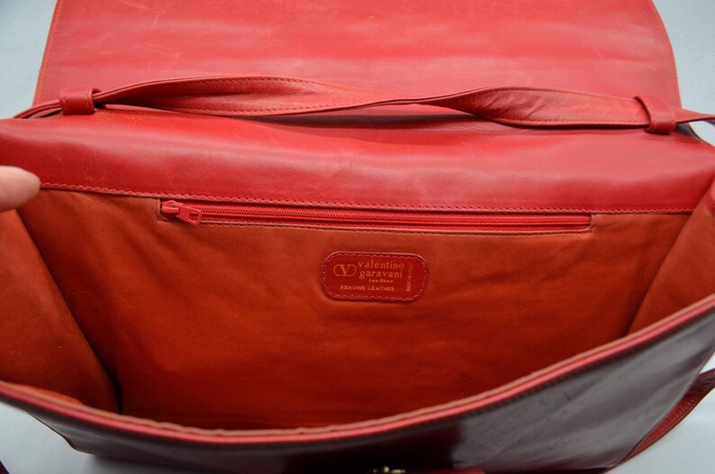 May include: Red leather handbag with a flap closure and a shoulder strap. The inside of the bag is lined with a red fabric and has a zippered pocket. The bag has a label that reads "Valentino Garavani Genuine Leather."