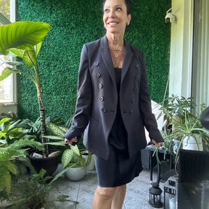 Puede incluir: Una mujer con un blazer negro de doble botonadura con una falda negra y un collar plateado. Está de pie frente a una pared verde.
