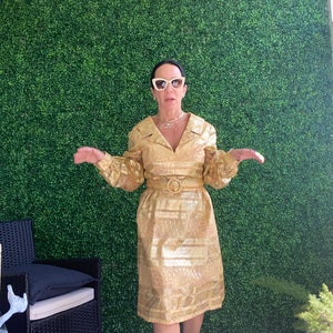 Op de afbeelding: Een vrouw die een gouden metallic jurk draagt met een riem en een witte zonnebril. Ze staat voor een groene muur.