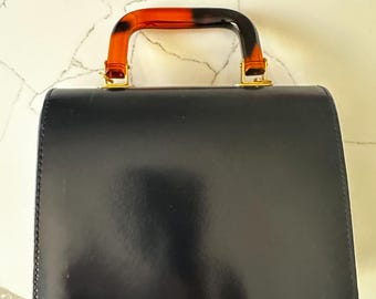 Puede incluir: Un bolso negro con un acabado brillante. El asa tiene un diseño curvo con un estampado de carey en tonos naranja y marrón. Los herrajes dorados aseguran el asa a la bolsa.