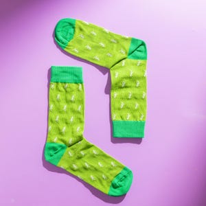 以下が含まれることがあります： 紫色の背景に、白い恐竜模様の緑色の靴下が2足。 靴下のかかとは明るい緑色で、つま先と袖口も緑色です。 恐竜の模様が靴下全体に繰り返されています。