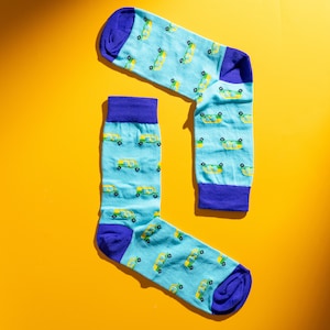 May include: A pair of novelty socks with a light blue background and a pattern of yellow and green vans. The socks have purple cuffs, heels, and toes. The socks are displayed on a yellow background.