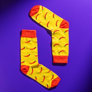 May include: A pair of yellow socks featuring a repeating pattern of red chilli peppers. The socks have red heels, toes, and cuffs. The background is a solid purple.