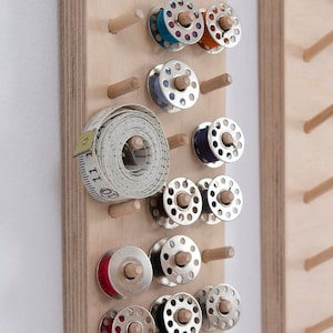 Può includere: Un organizzatore da parete in legno con pioli per riporre bobine di filo. L'organizzatore contiene una varietà di bobine di filo di diversi colori, tra cui bianco, rosso, blu e verde. Un metro a nastro è anche appeso all'organizzatore.