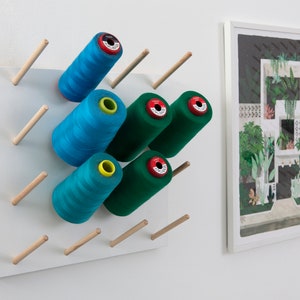 May include: A white pegboard with wooden dowels holds spools of teal and green thread. The thread spools have red, yellow, and white labels. A framed artwork with a white border is on the right.