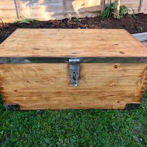 May include: A rectangular wooden storage chest with a light brown finish. The chest has metal corner protectors and a metal clasp. The edges are reinforced with metal. The chest is sitting on green grass.